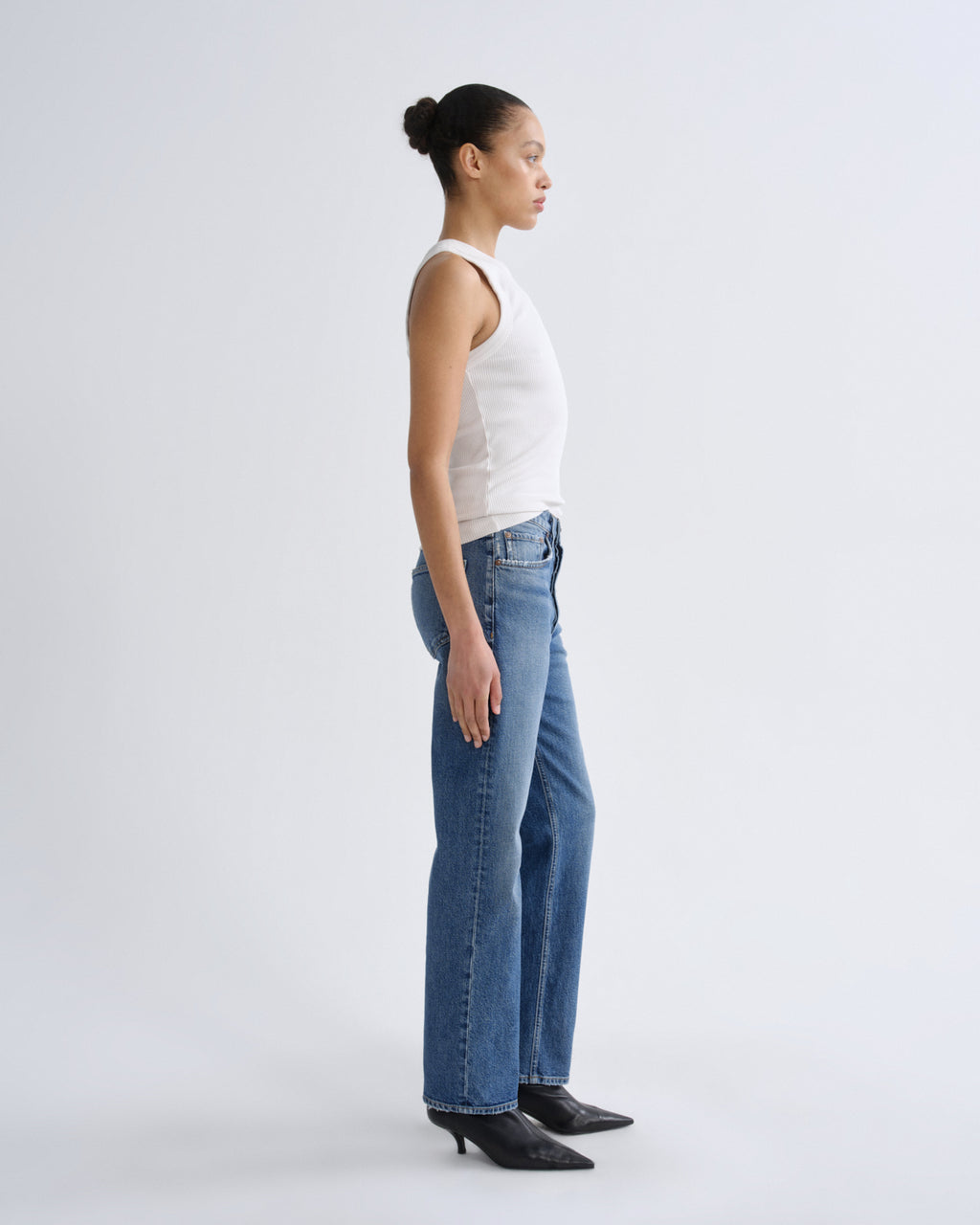Woman wearing a white tank top and blue jeans standing against a plain background