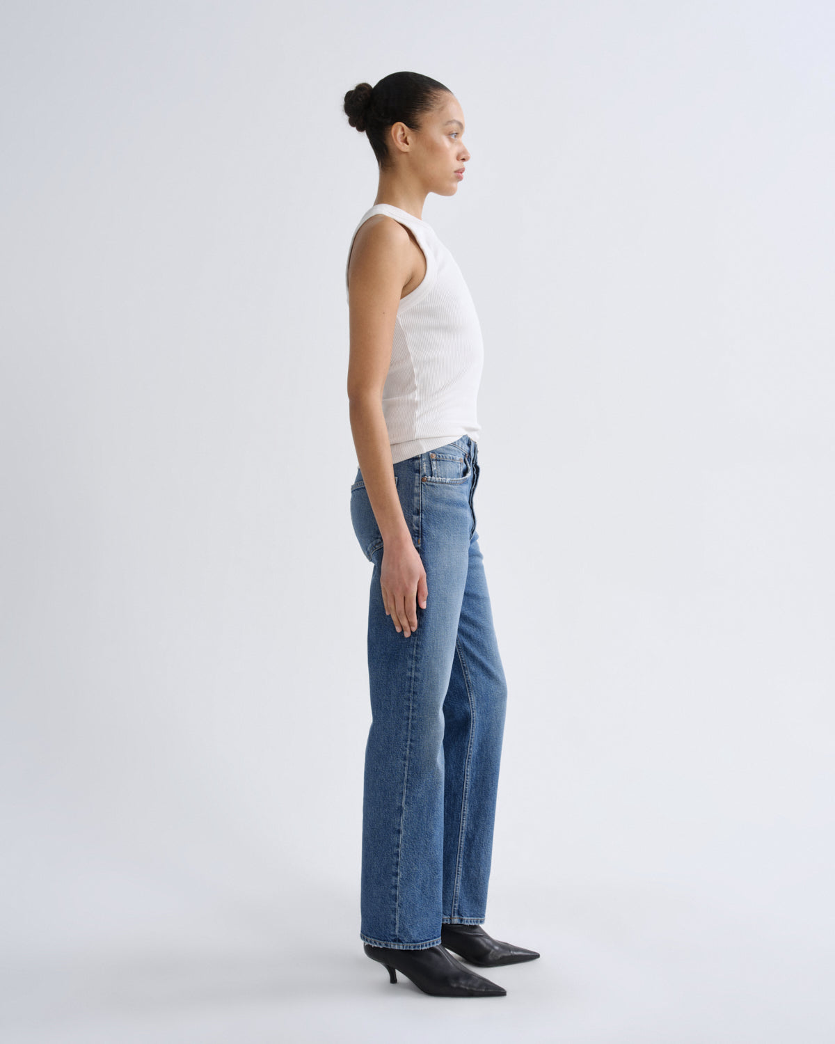 Woman wearing a white tank top and blue jeans standing against a plain background