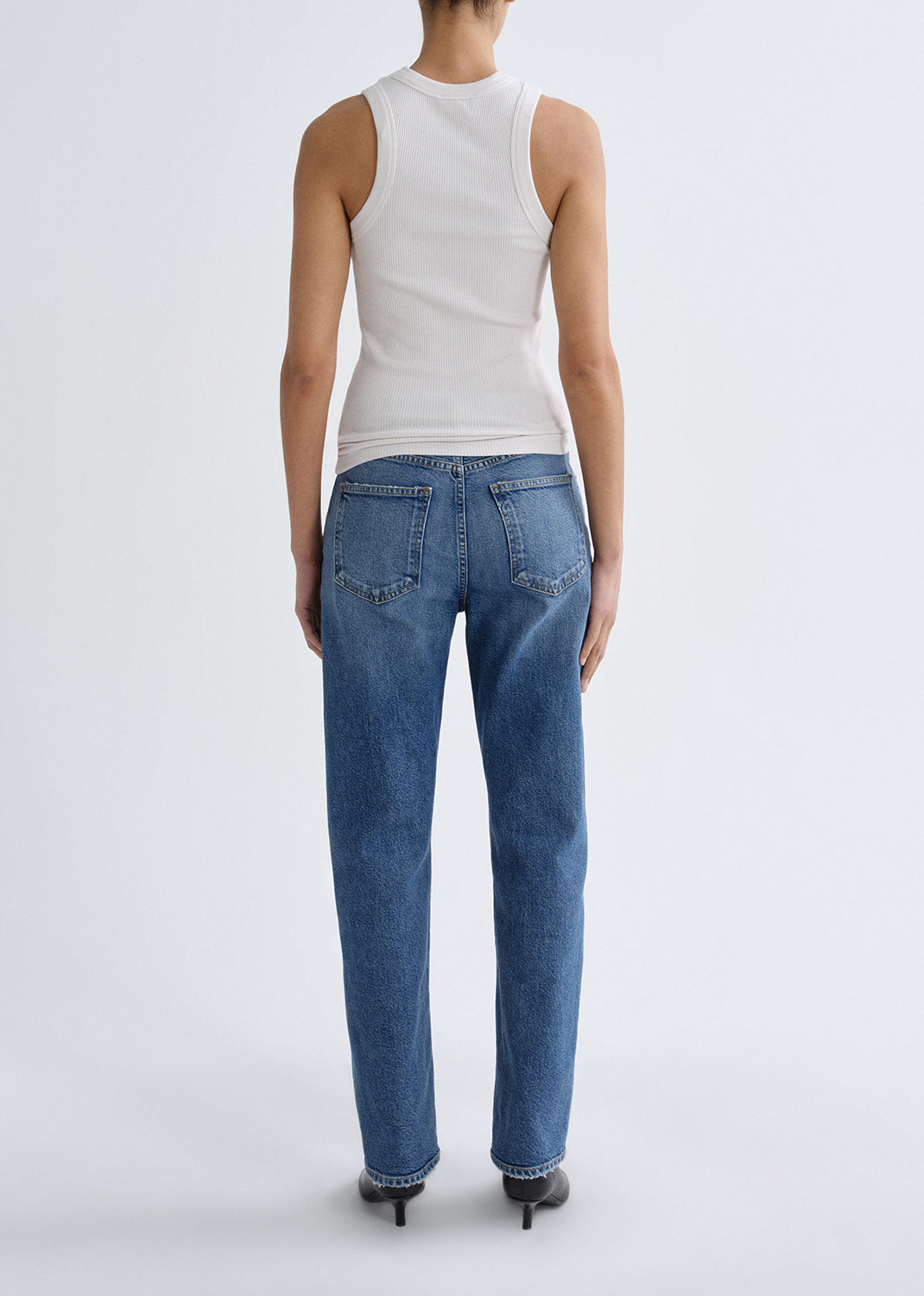 A model shown from the back wearing a white sleeveless top and blue straight-leg jeans, standing against a grey background.