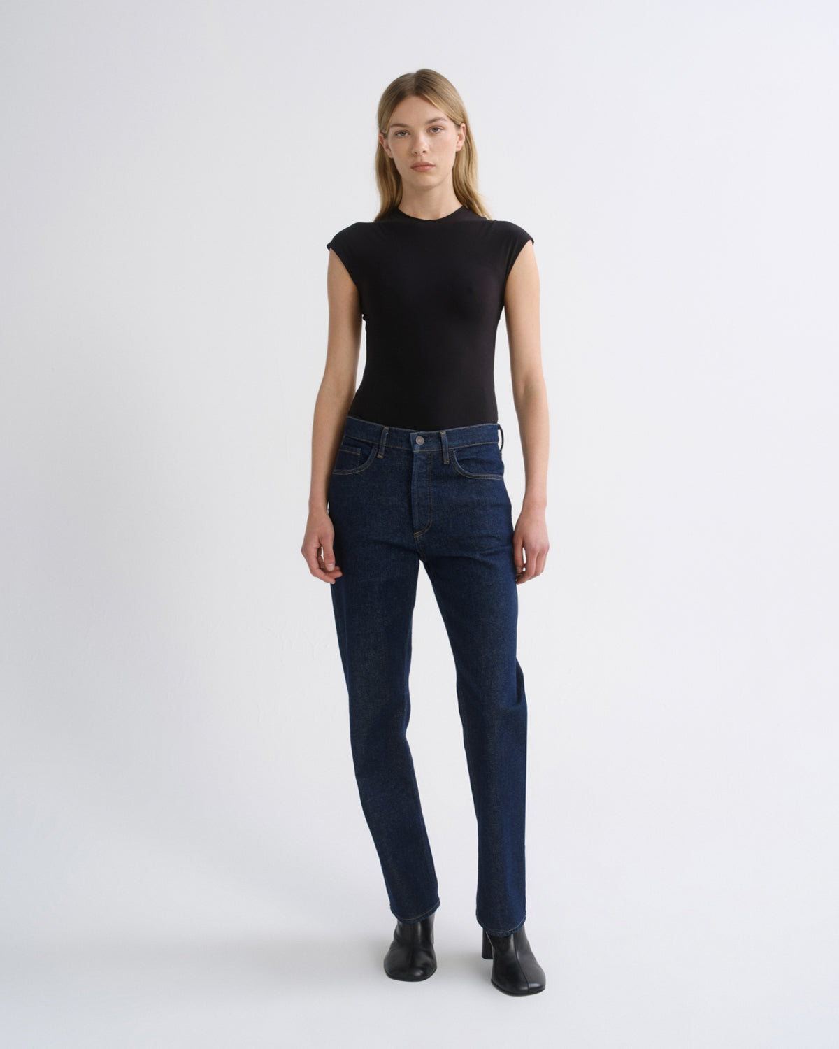 Woman wearing a black cap sleeve top with dark indigo straight leg jeans on a grey background.
