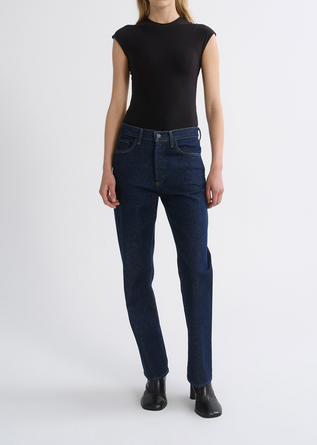A model wearing dark blue straight-leg jeans, a black sleeveless top, and black boots, standing against a grey background.