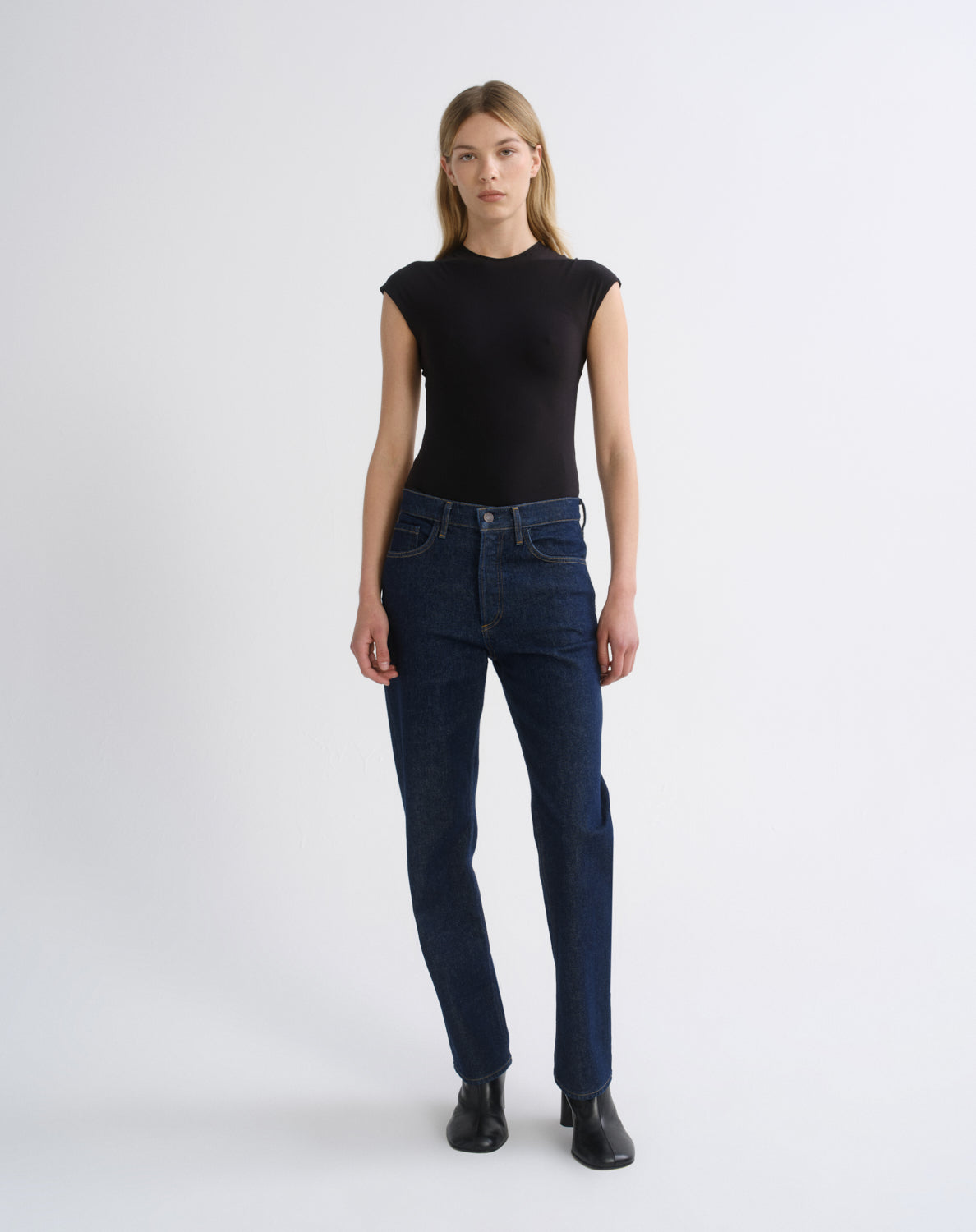 A model stands against a light gray studio background wearing a fitted black short-sleeve top, dark blue straight-leg jeans, and black ankle boots.