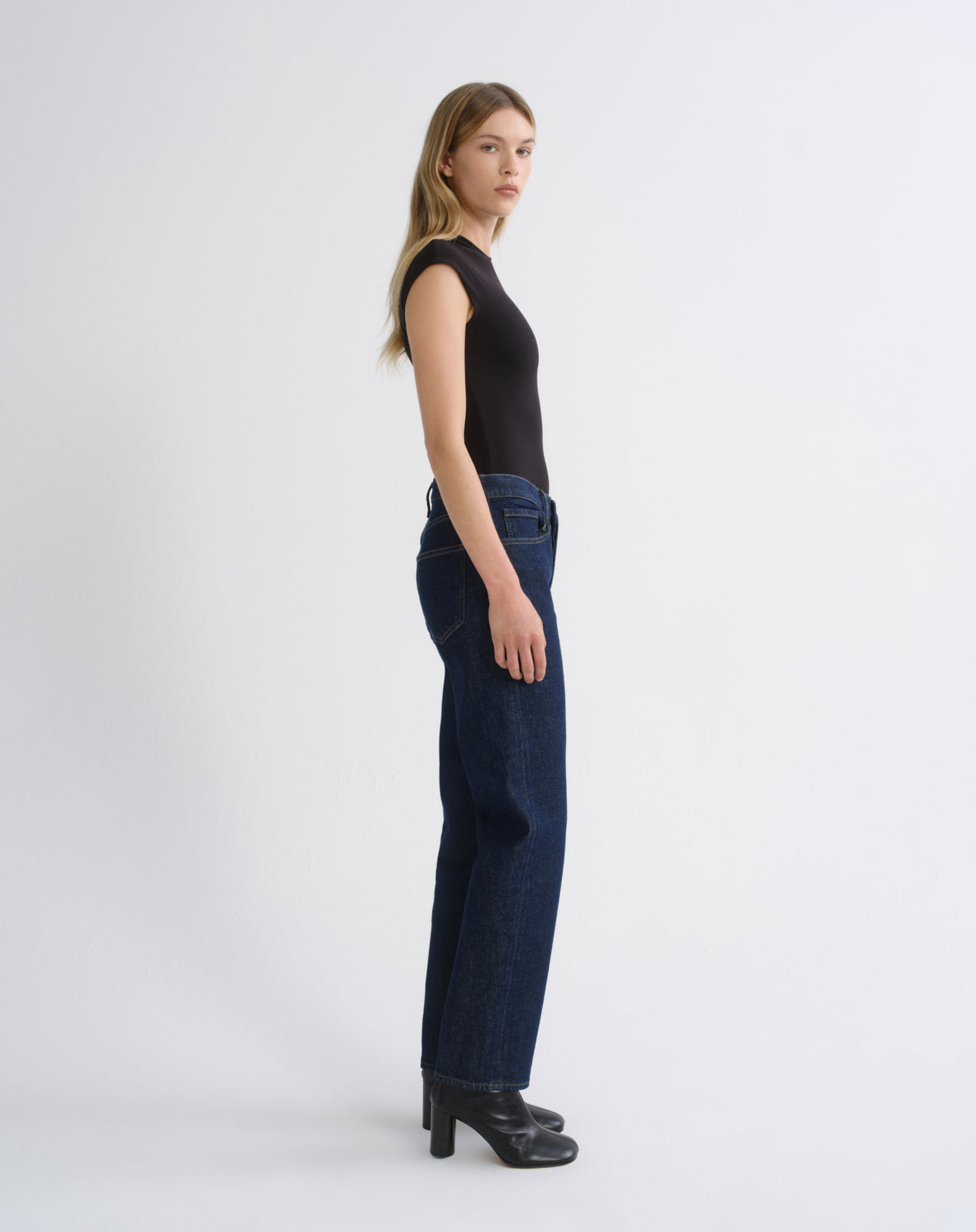 A woman stands in profile wearing a black sleeveless top, dark blue wide-leg jeans, and black heeled boots against a plain light gray studio background.
