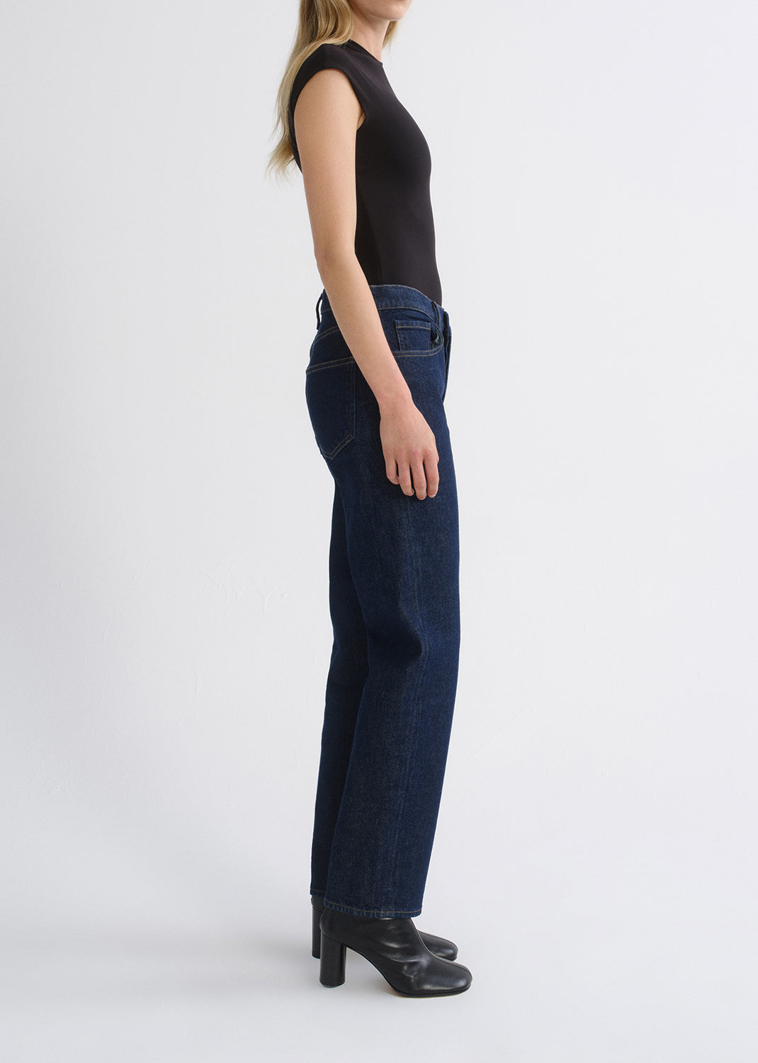 A side-profile view of a model wearing dark blue straight-leg denim jeans, a black sleeveless top, and black heeled boots against a grey background.