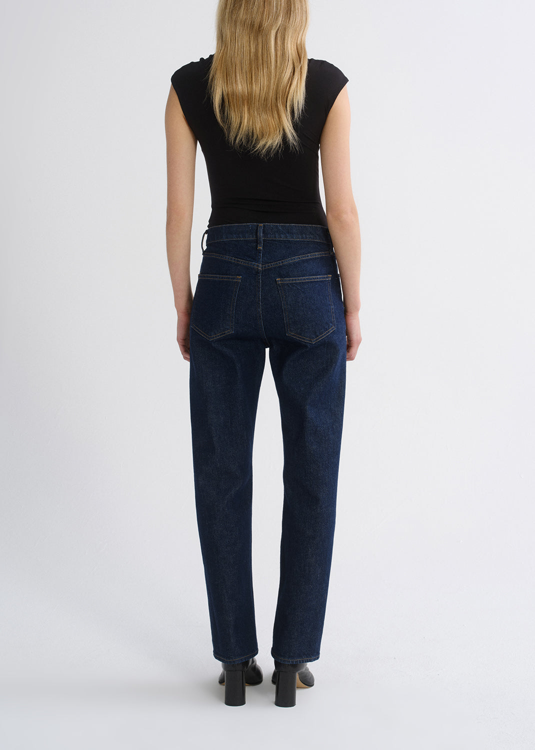 A woman viewed from the back wearing dark blue straight-leg jeans, a black sleeveless top, and black heels against a grey background.