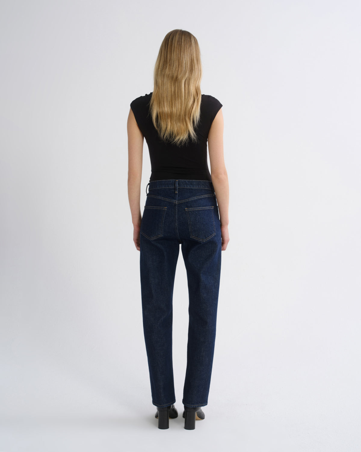 A woman viewed from behind wearing dark blue straight-leg jeans, a black sleeveless top, and black heels against a plain light gray studio background.