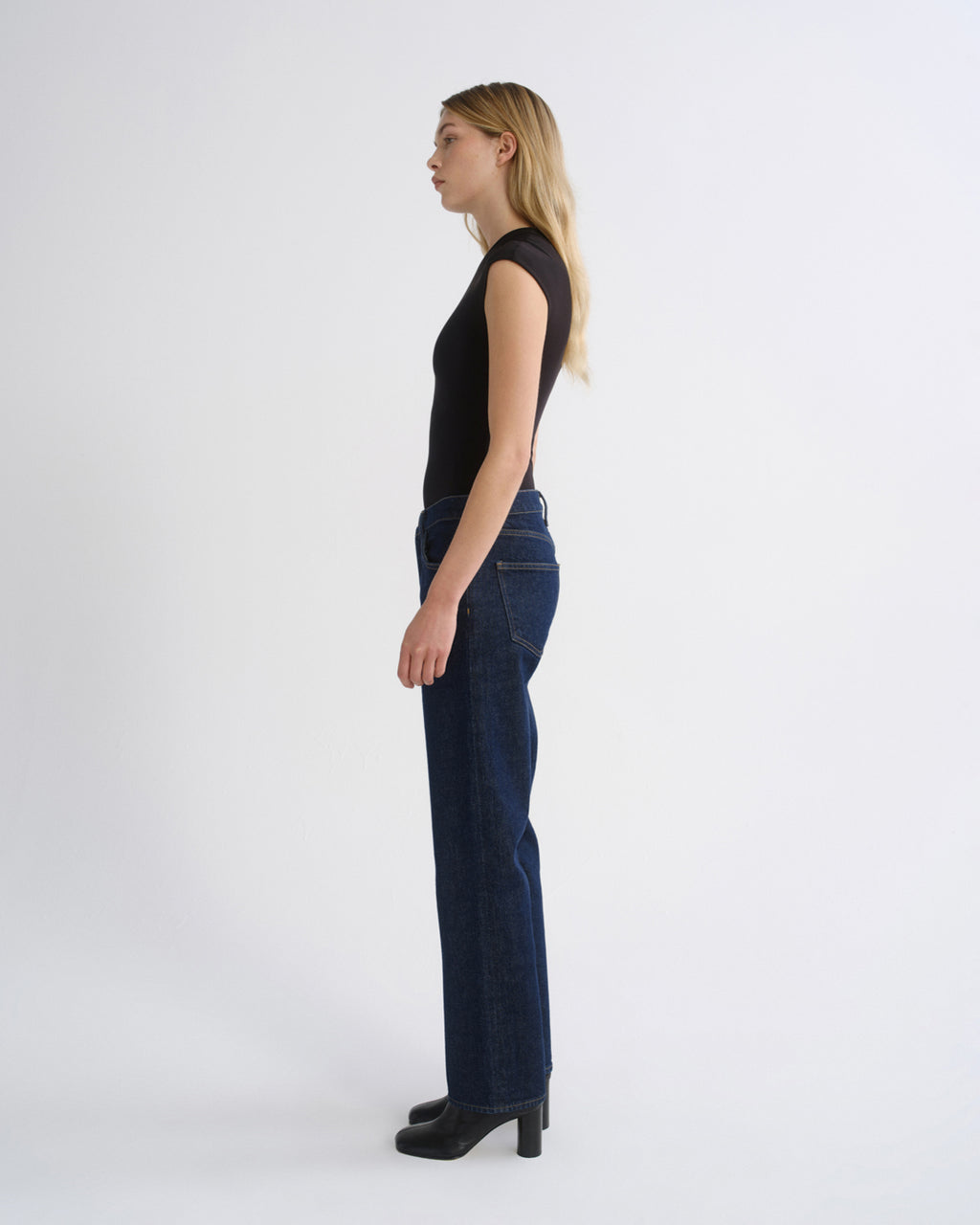 Woman wearing a black cap sleeve top with dark indigo straight leg jeans viewed from the side on a grey background.