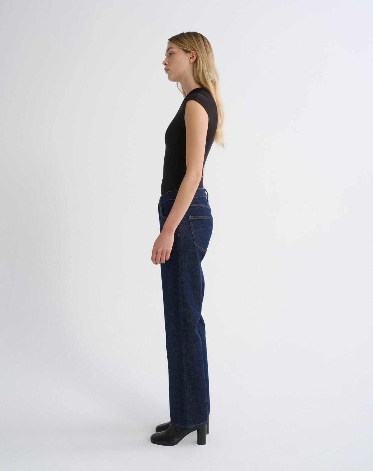A woman stands in profile wearing a black sleeveless top and dark blue straight-leg jeans with black heels, photographed against a clean light gray studio background.