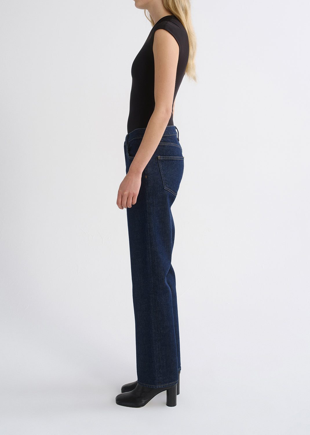 A side-profile studio shot of a model wearing dark blue straight-leg jeans, a black sleeveless top, and black heeled boots against a grey background.
