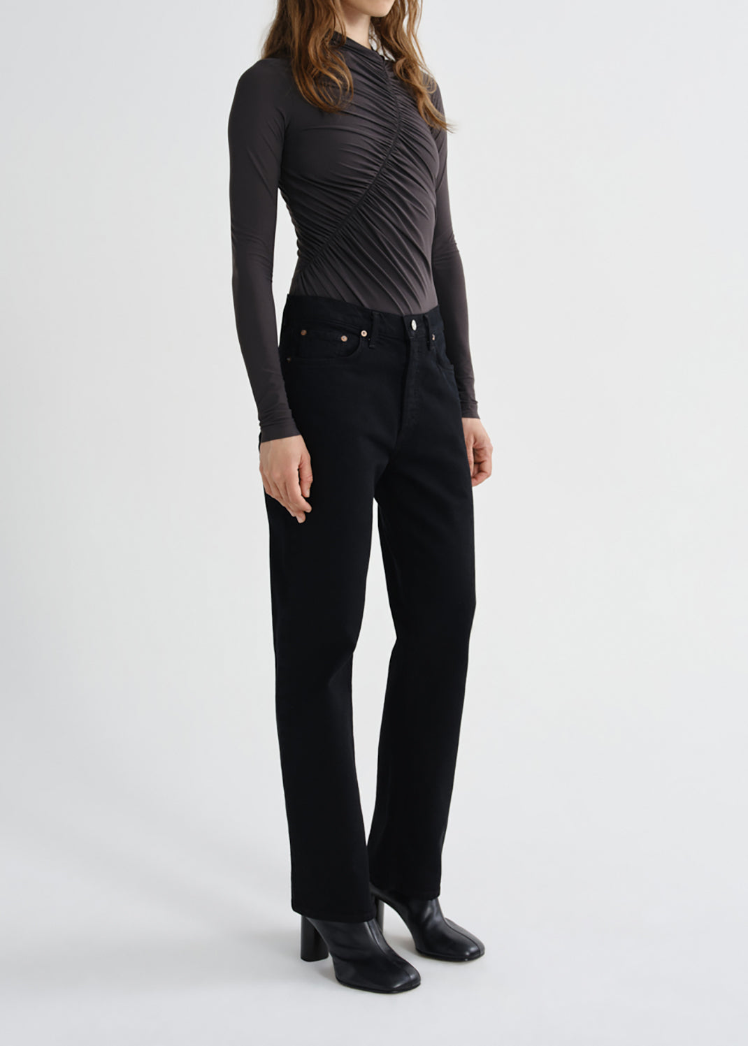 A model wearing a dark gray ruched long-sleeve top and black wide-leg jeans with black leather boots, standing against a grey background.