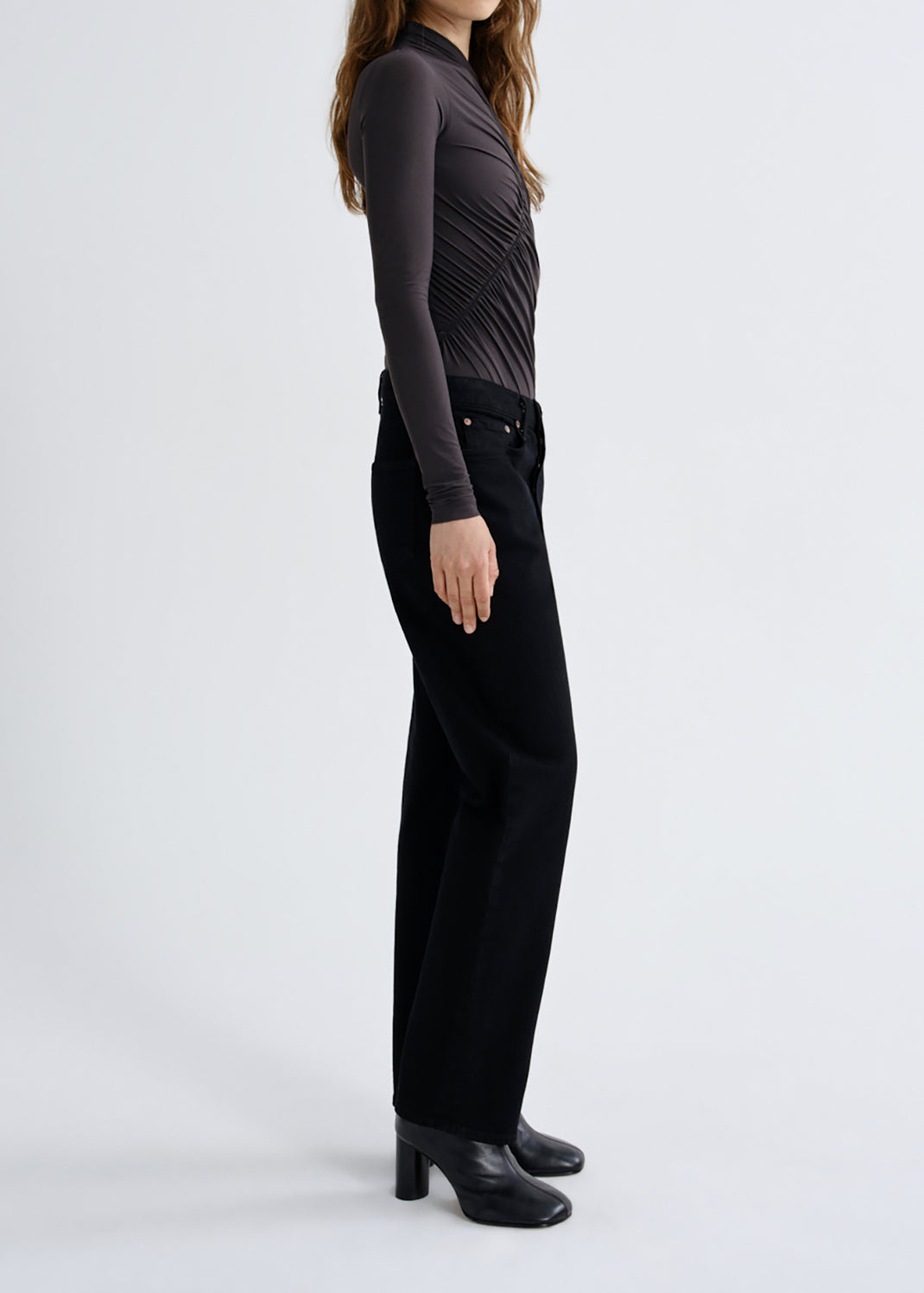 A model shown from the side wearing a dark charcoal gray long-sleeve top with a ruched, pleated texture and black wide-leg trousers, paired with black leather ankle boots, against a grey background.