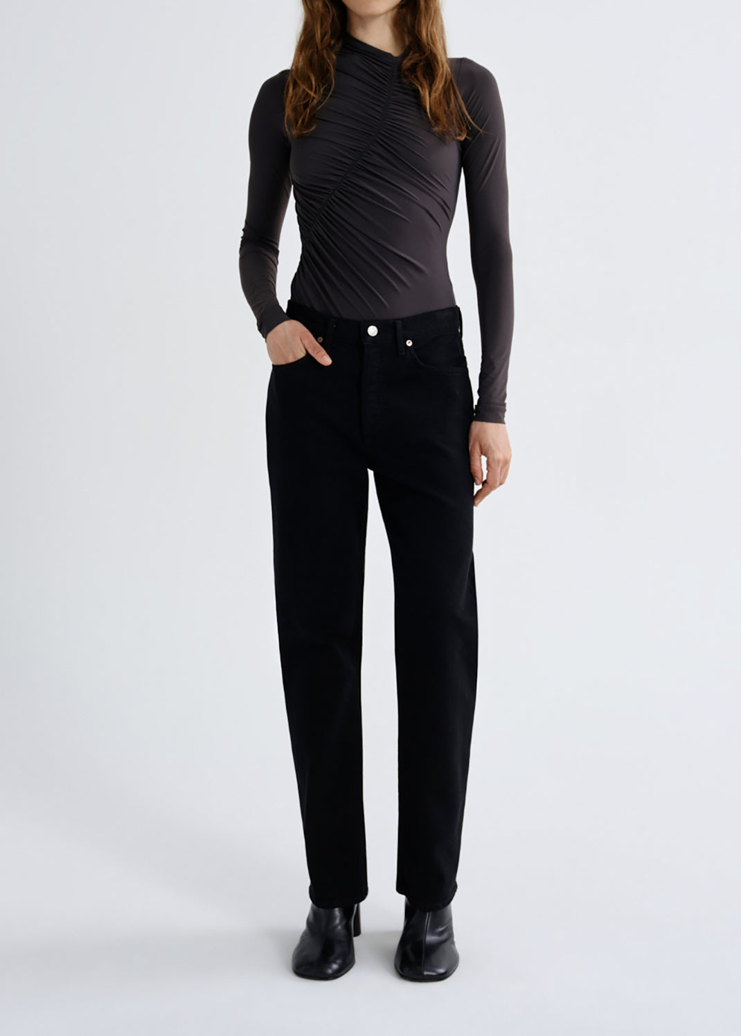 A model wearing a black long-sleeve ruched mock-neck top and black trousers, standing against a grey background.