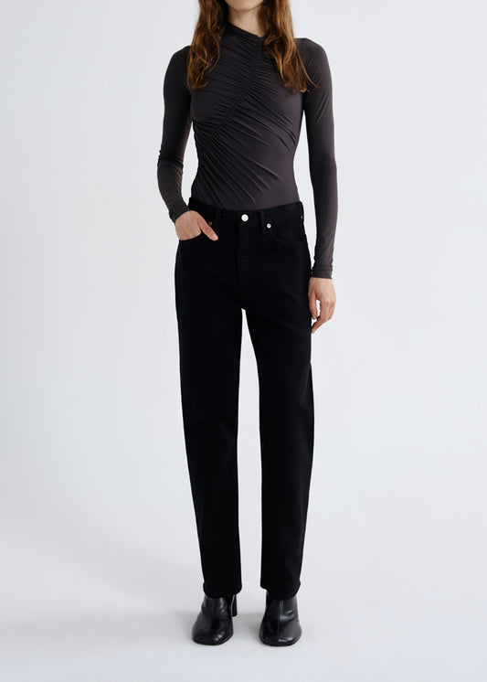 A model wearing a black long-sleeve ruched mock-neck top and black trousers, standing against a grey background.