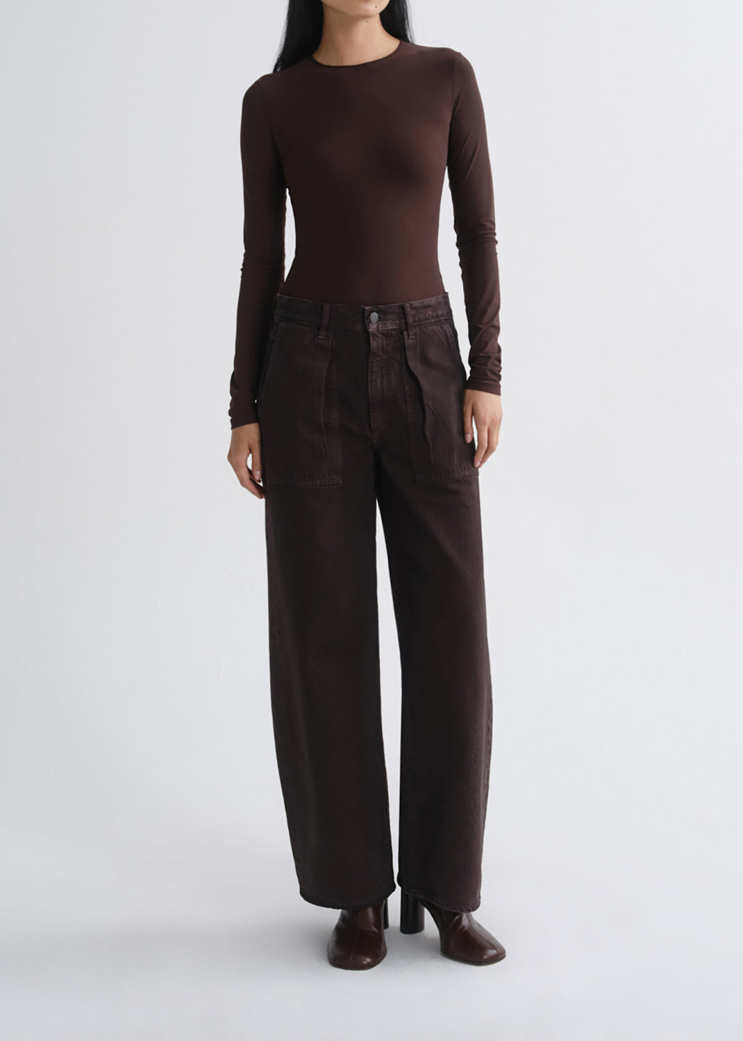A model wearing a dark brown long-sleeve crewneck top and matching dark brown wide-leg trousers, standing against a grey background.