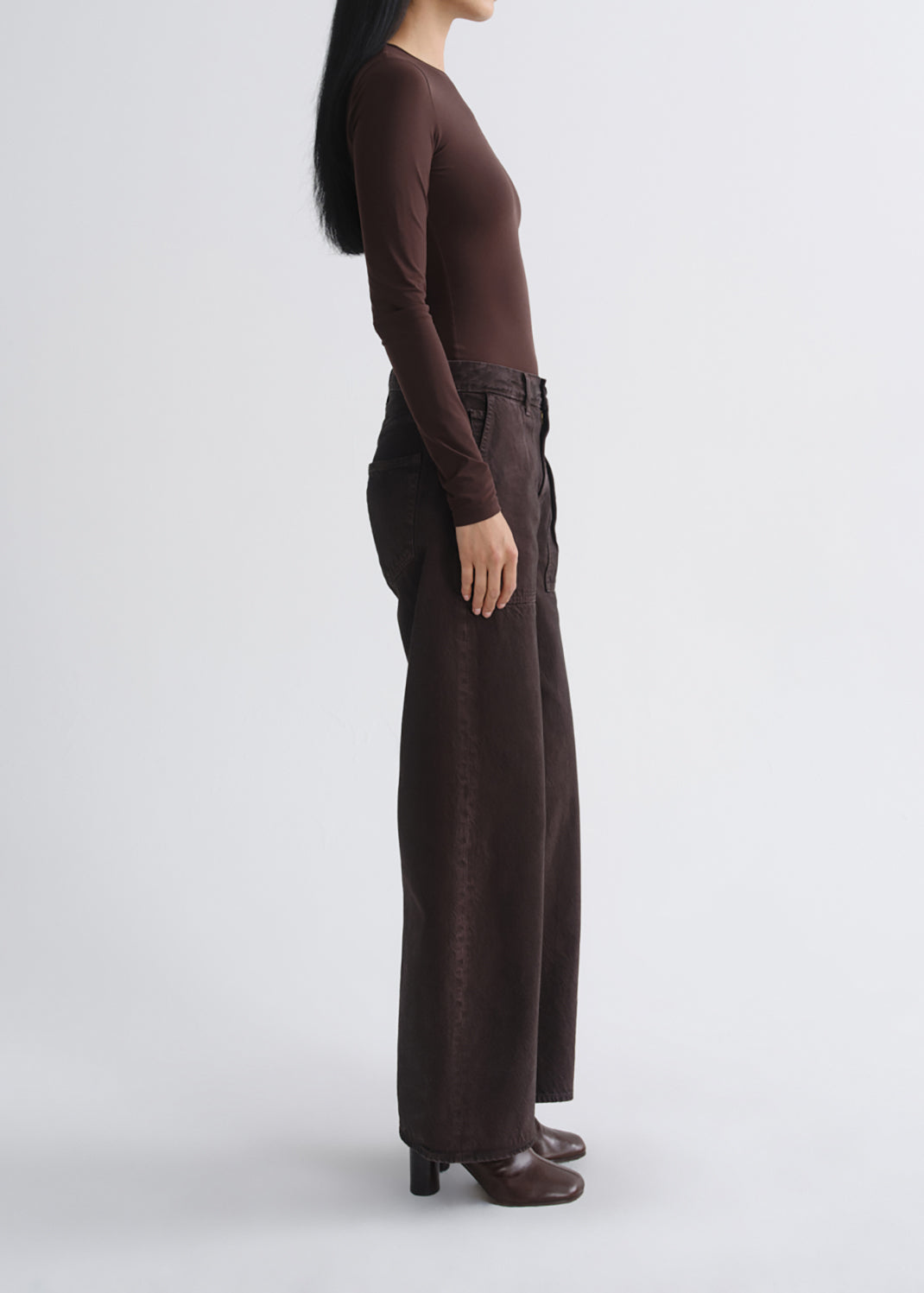 A model shown in profile wearing a dark brown long-sleeve top and matching dark brown wide-leg trousers, paired with dark brown heeled boots, against a grey background.
