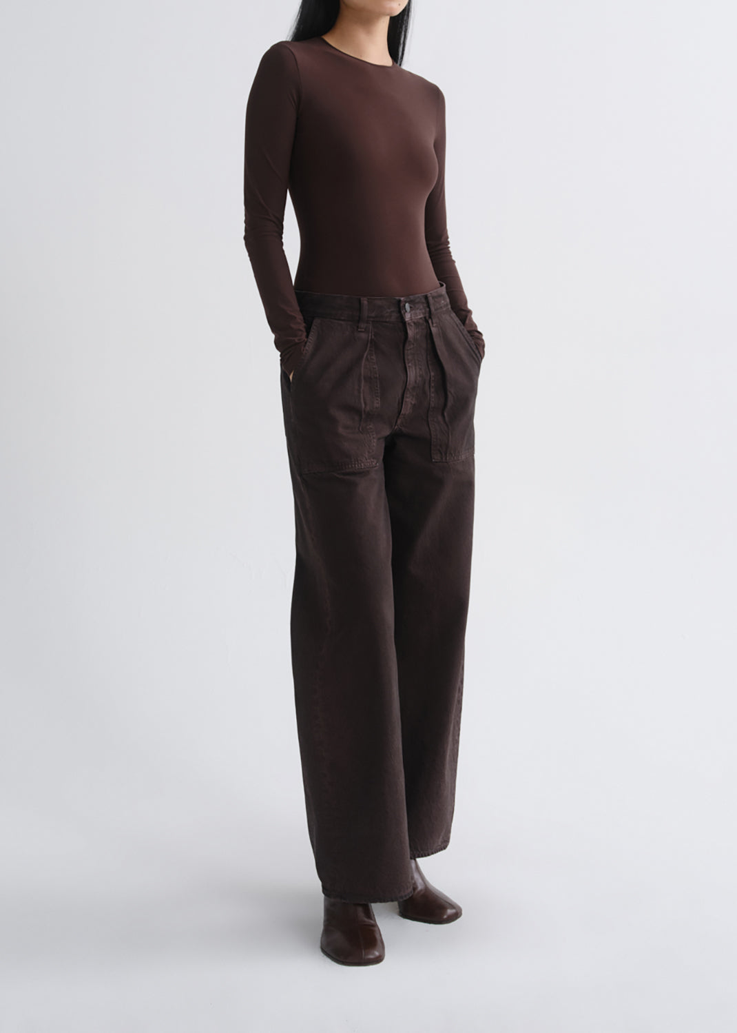 A model wearing a dark brown long-sleeve crewneck top and matching dark brown wide-leg trousers, standing against a grey background.