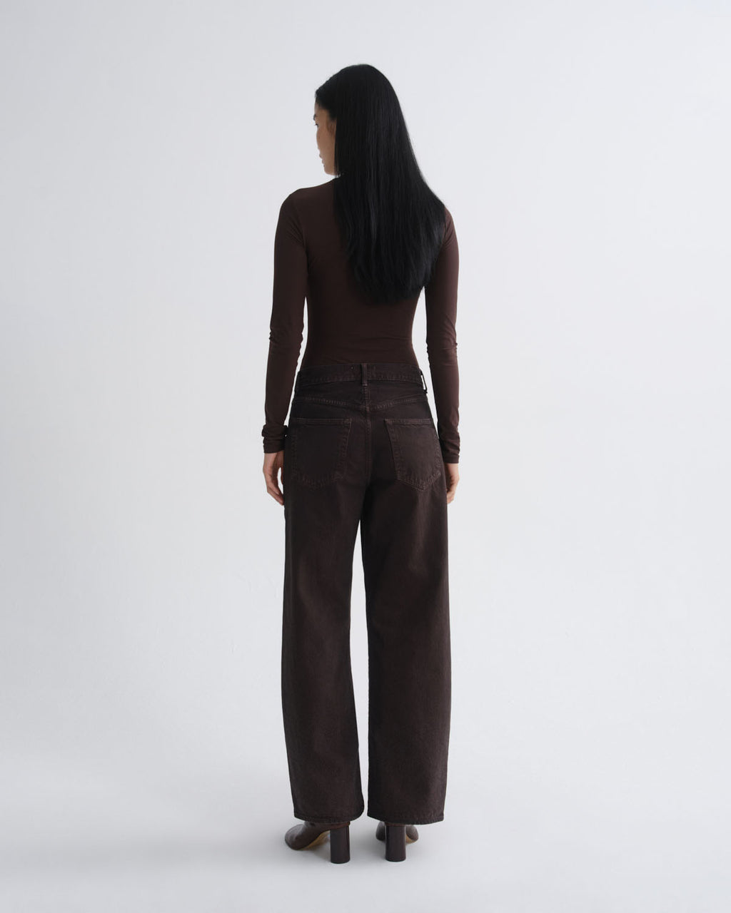 A woman viewed from behind wearing a dark brown long-sleeve top and matching dark brown wide-leg trousers, standing against a light gray studio background.