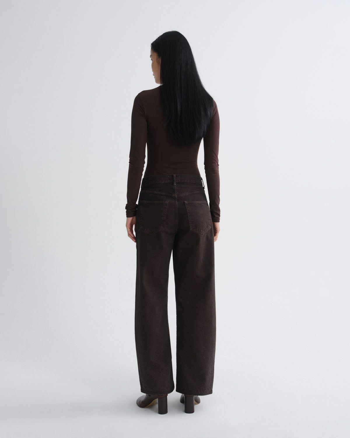 A woman viewed from behind wearing a dark brown long-sleeve top and matching dark brown wide-leg trousers, standing against a light gray studio background.