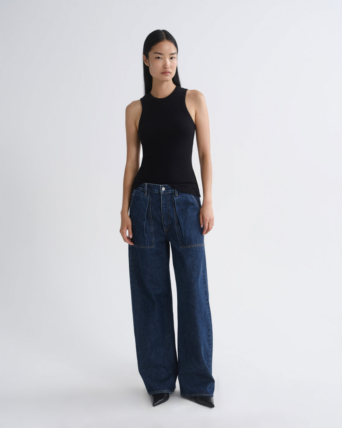 A model stands against a light gray studio background wearing a black sleeveless ribbed top and dark blue wide-leg denim jeans.