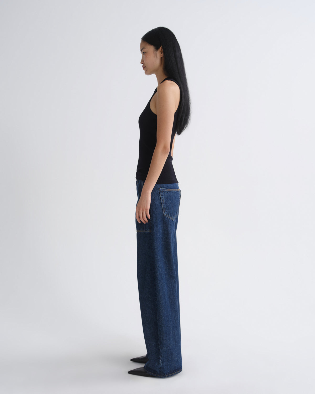 A woman stands in profile wearing a black sleeveless top and wide-leg dark blue jeans against a plain light gray studio background.