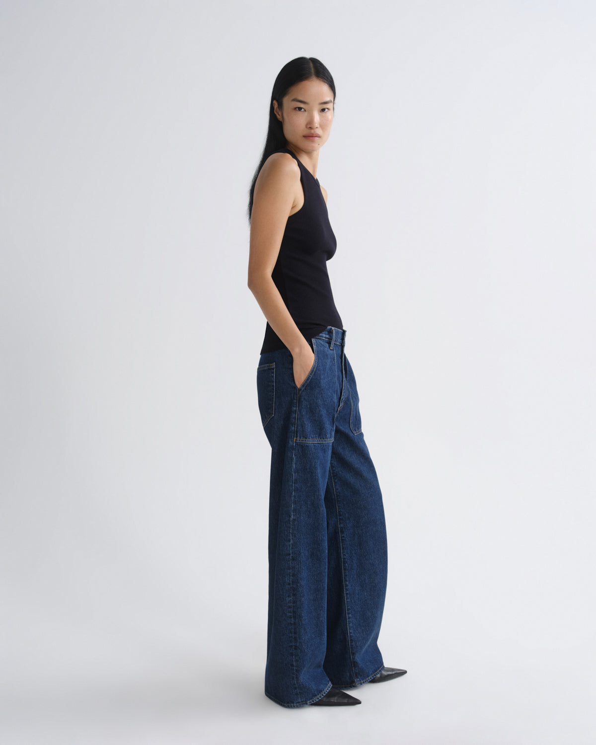 A woman stands in profile wearing a black sleeveless top and dark blue wide-leg jeans against a plain light gray studio background.