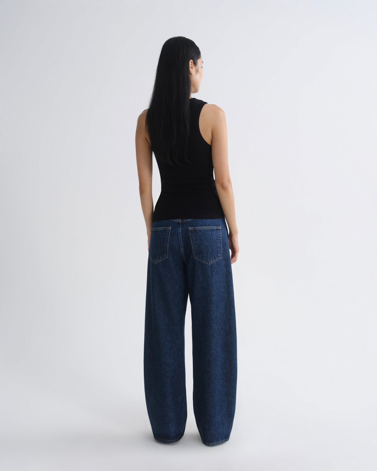 A woman is shown from the back wearing a black sleeveless top and dark blue wide-leg jeans against a plain light gray studio background.