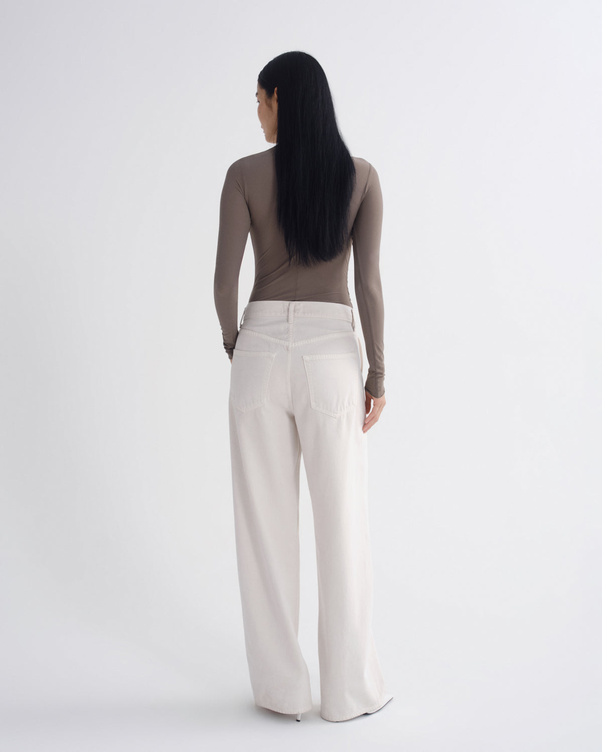 A woman with long black hair is shown from behind wearing a taupe long-sleeve top and off-white wide-leg trousers against a plain white studio background.