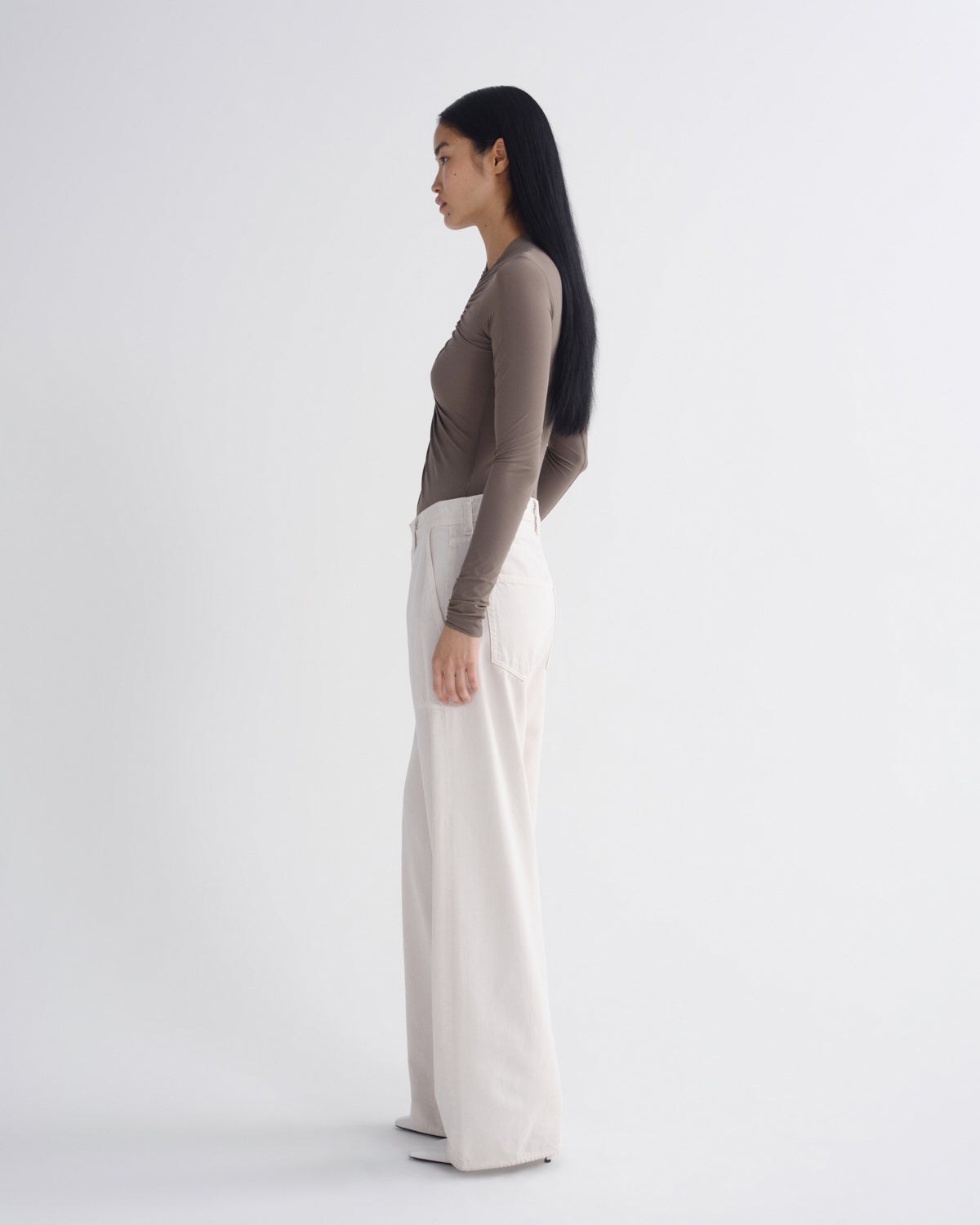 A woman stands in profile against a plain light gray studio background, wearing a fitted taupe-brown long-sleeve top and off-white wide-leg pants.