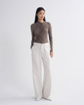 A model stands against a light gray studio background wearing a taupe-brown long-sleeve top with diagonal ruching and pleats, paired with off-white wide-leg trousers.