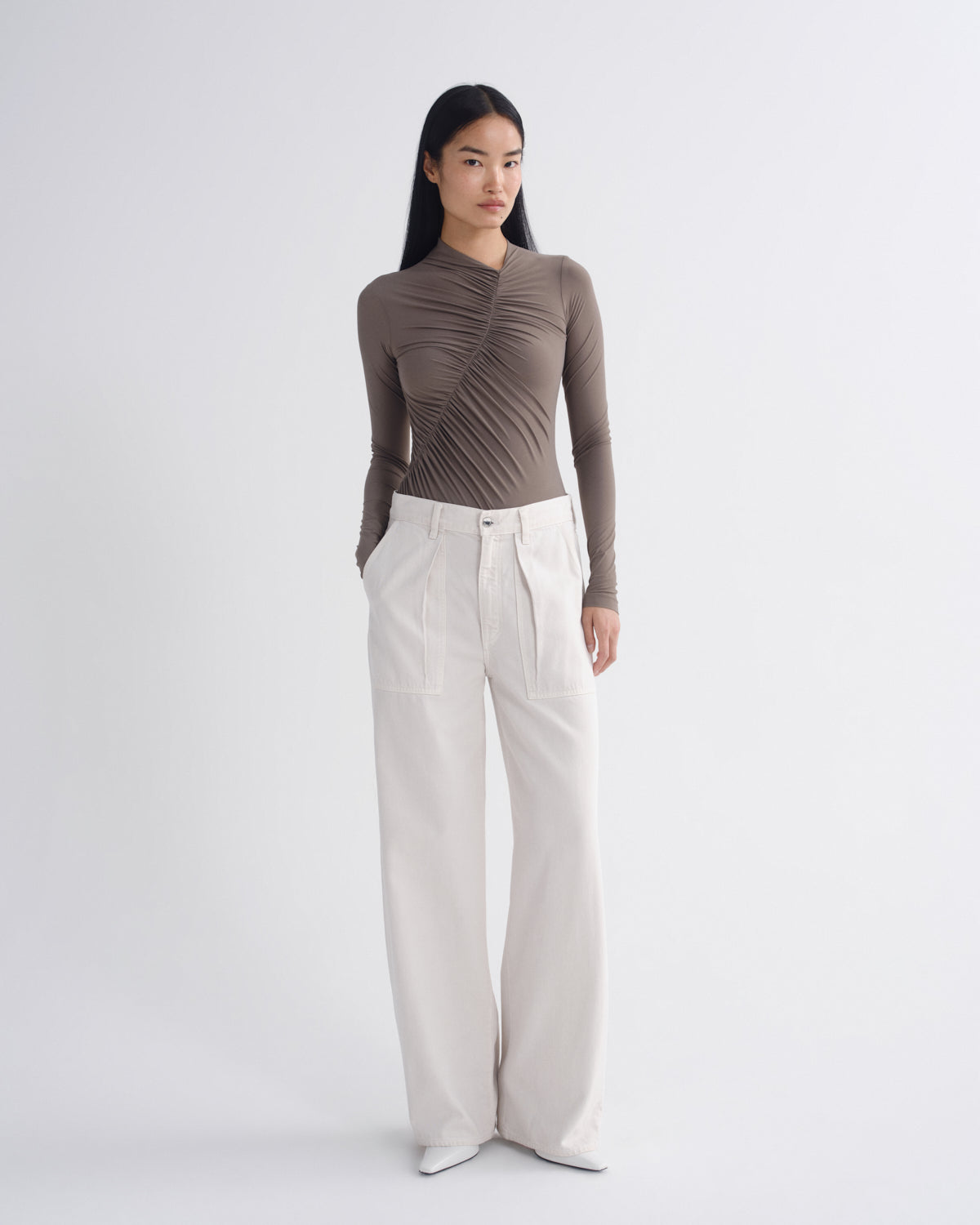 A model stands against a light gray studio background wearing a taupe-brown long-sleeve top with diagonal ruching and pleats, paired with off-white wide-leg trousers.