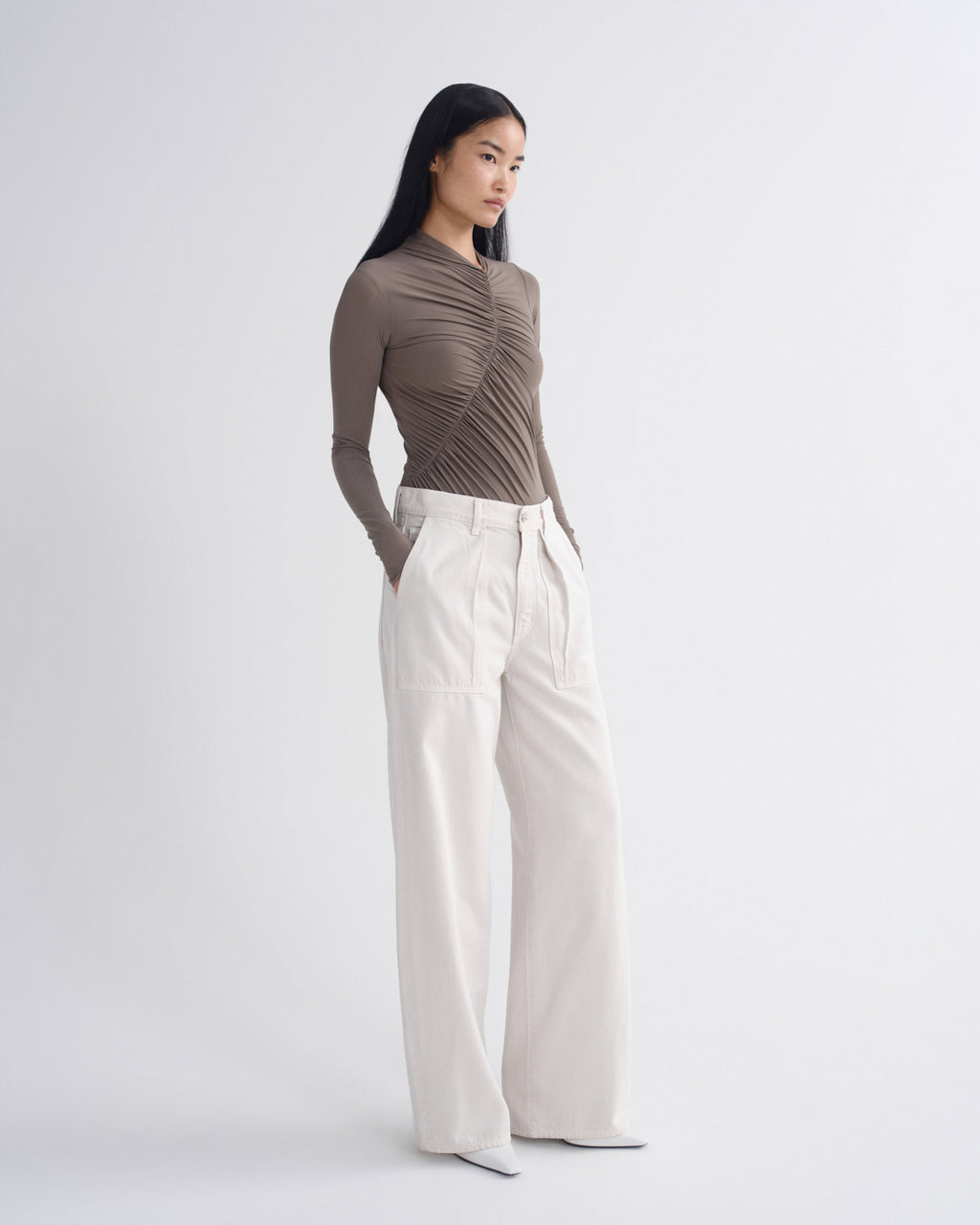 A model stands in a studio wearing a taupe-brown long-sleeve top with dramatic vertical pleats and high-waisted off-white wide-leg trousers.
