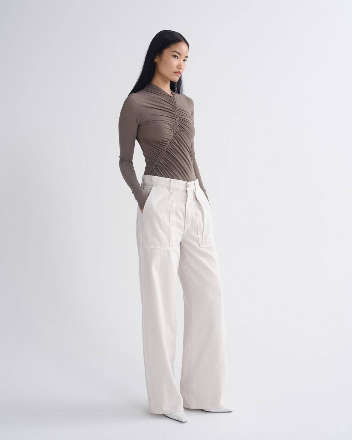 A model stands in a studio wearing a taupe-brown long-sleeve top with dramatic vertical pleats and high-waisted off-white wide-leg trousers.