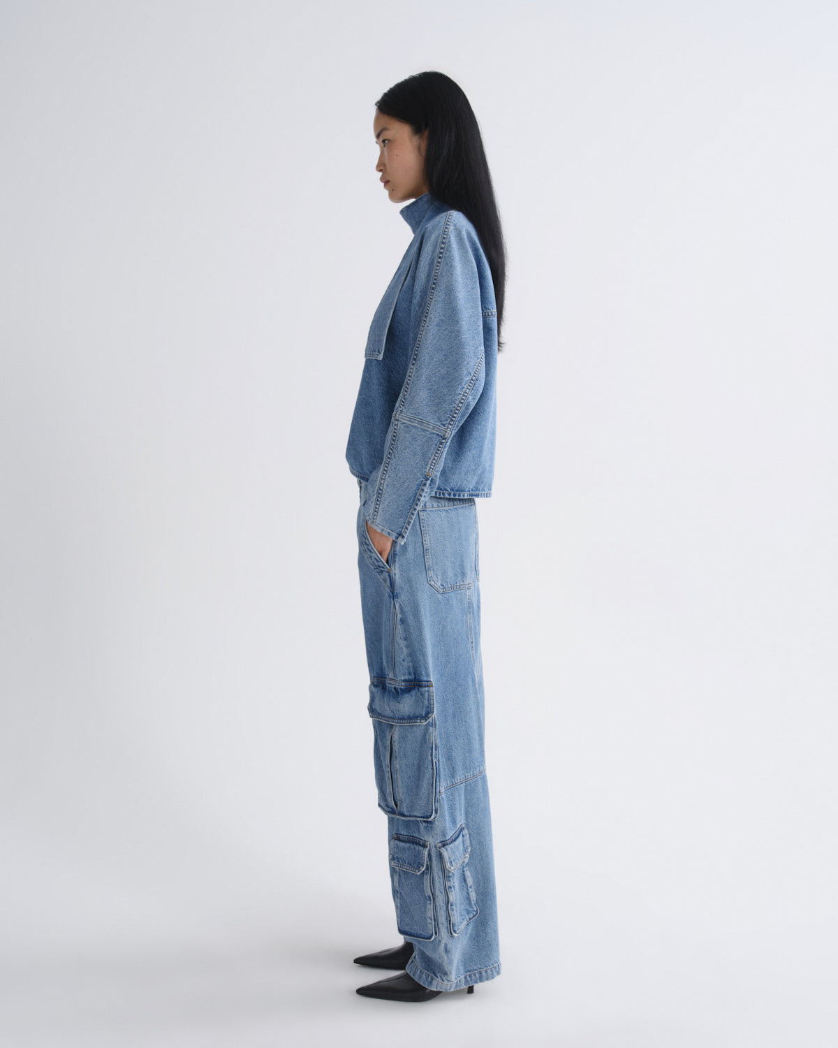 A model in side profile wearing a light blue denim jacket and matching wide-leg cargo pants against a plain light gray studio background.
