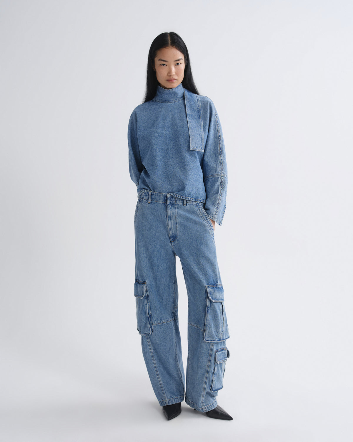 A model stands against a light gray studio background wearing a matching blue denim set consisting of a high-neck long-sleeve top and wide-leg cargo pants.