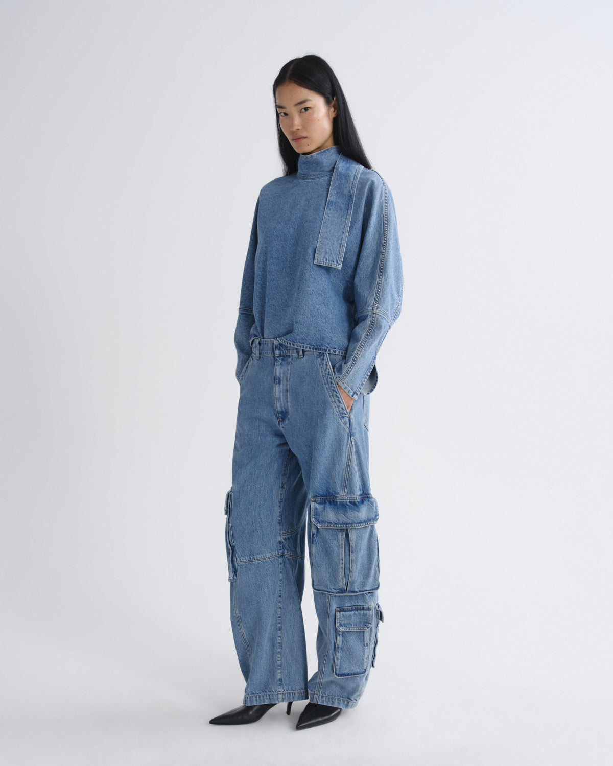 A model wearing a matching light blue denim set consisting of a long-sleeve mock-neck top and wide-leg cargo pants with multiple flap pockets, paired with black pointed-toe heels, against a plain light gray studio background.