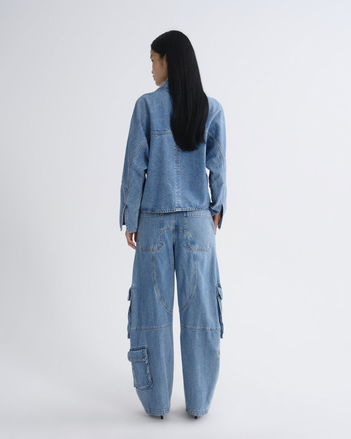 A model viewed from the back wearing a light blue denim set consisting of a cropped long-sleeve shirt and matching wide-leg cargo pants, shot against a plain light gray studio background.