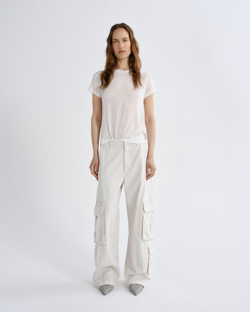 A woman stands against a light gray studio background wearing a loose white short-sleeve T-shirt and matching white wide-leg cargo pants with gray pointed shoes.