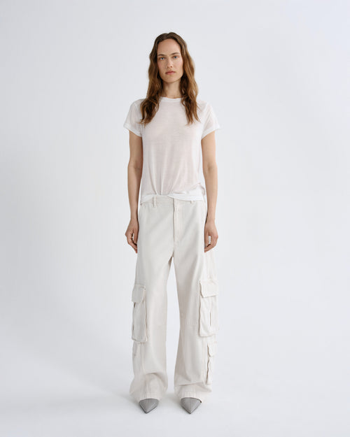 A woman stands against a light gray studio background wearing a loose white short-sleeve T-shirt and matching white wide-leg cargo pants with gray pointed shoes.