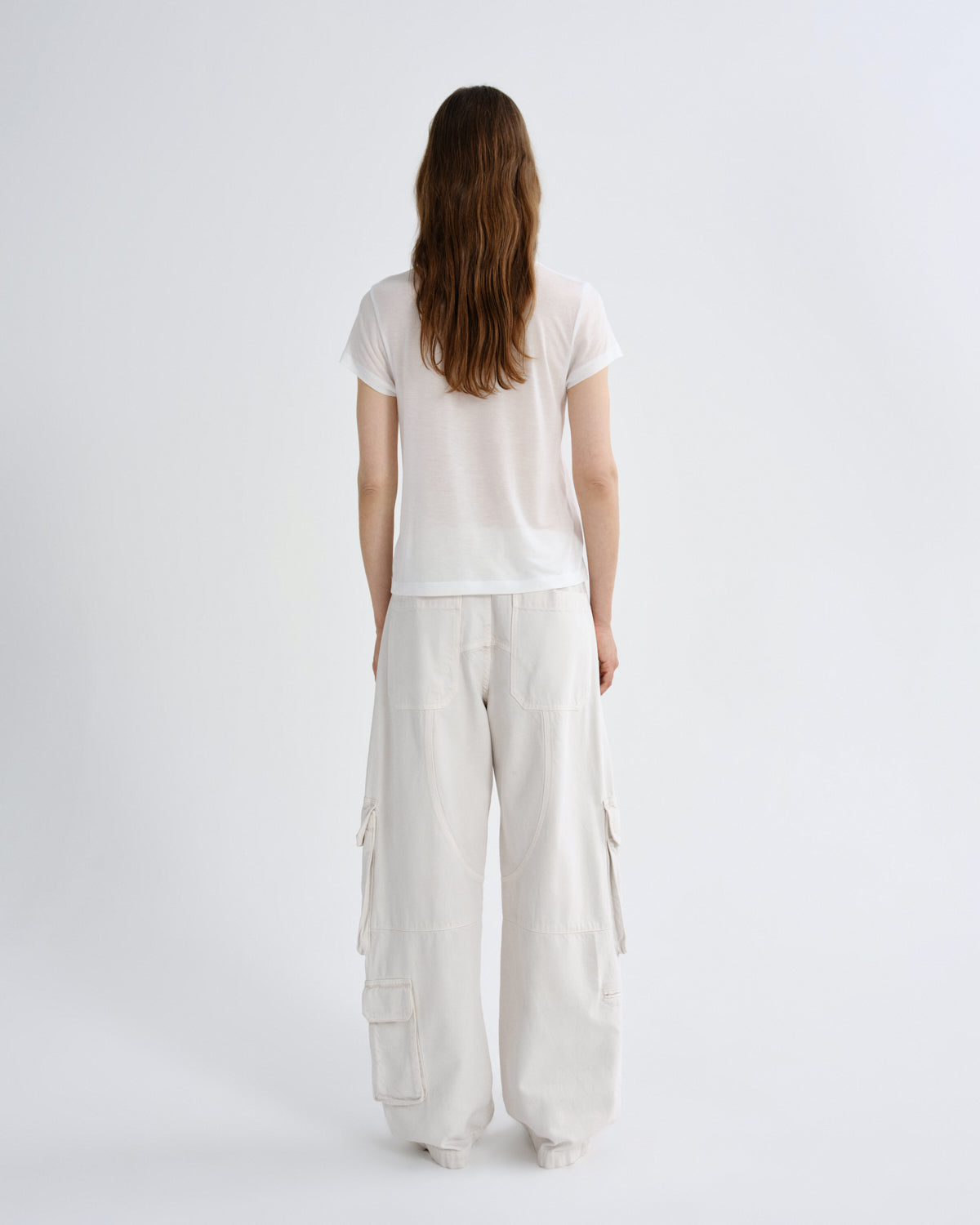 A woman viewed from behind wearing a white short-sleeve t-shirt and off-white wide-leg cargo pants against a plain white studio background.