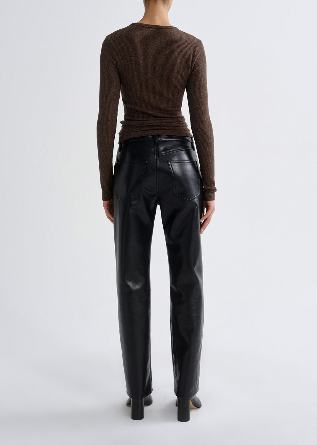 Person wearing a brown fitted long sleeve top with black straight leg leather pants viewed from the back on a grey background