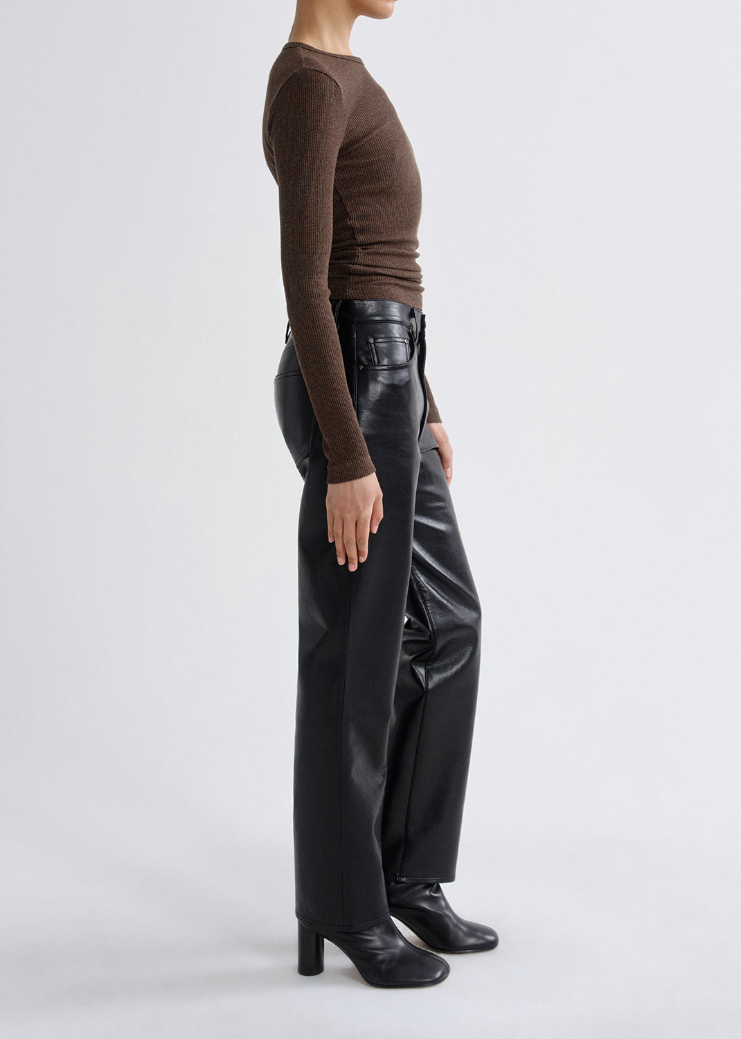 Person wearing a brown fitted long sleeve top with black straight leg leather pants viewed from the side on a grey background