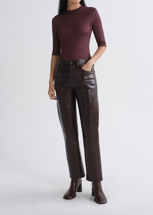 A model wearing a dark burgundy mock-neck knit top, dark brown leather pants, and dark brown heeled ankle boots, standing against a grey background.