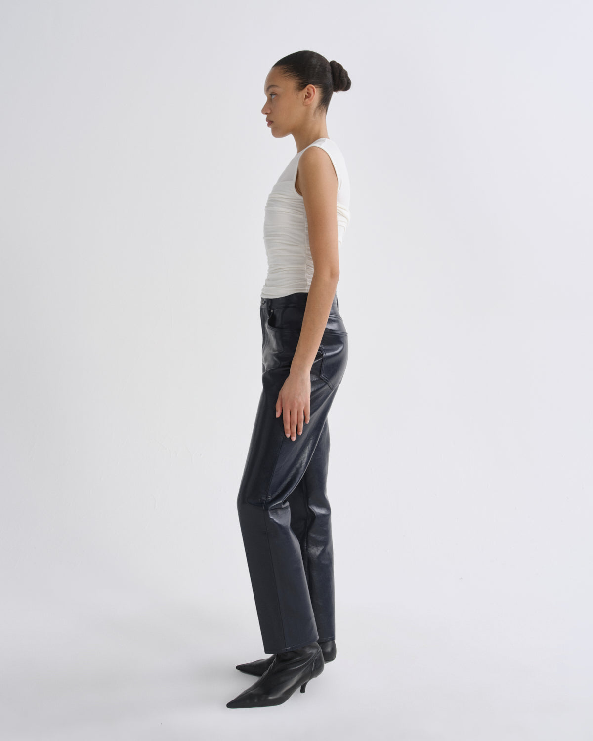 A model is shown in profile wearing a white ribbed sleeveless top, dark navy wide-leg trousers, and black pointed-toe ankle boots against a plain light gray studio background.