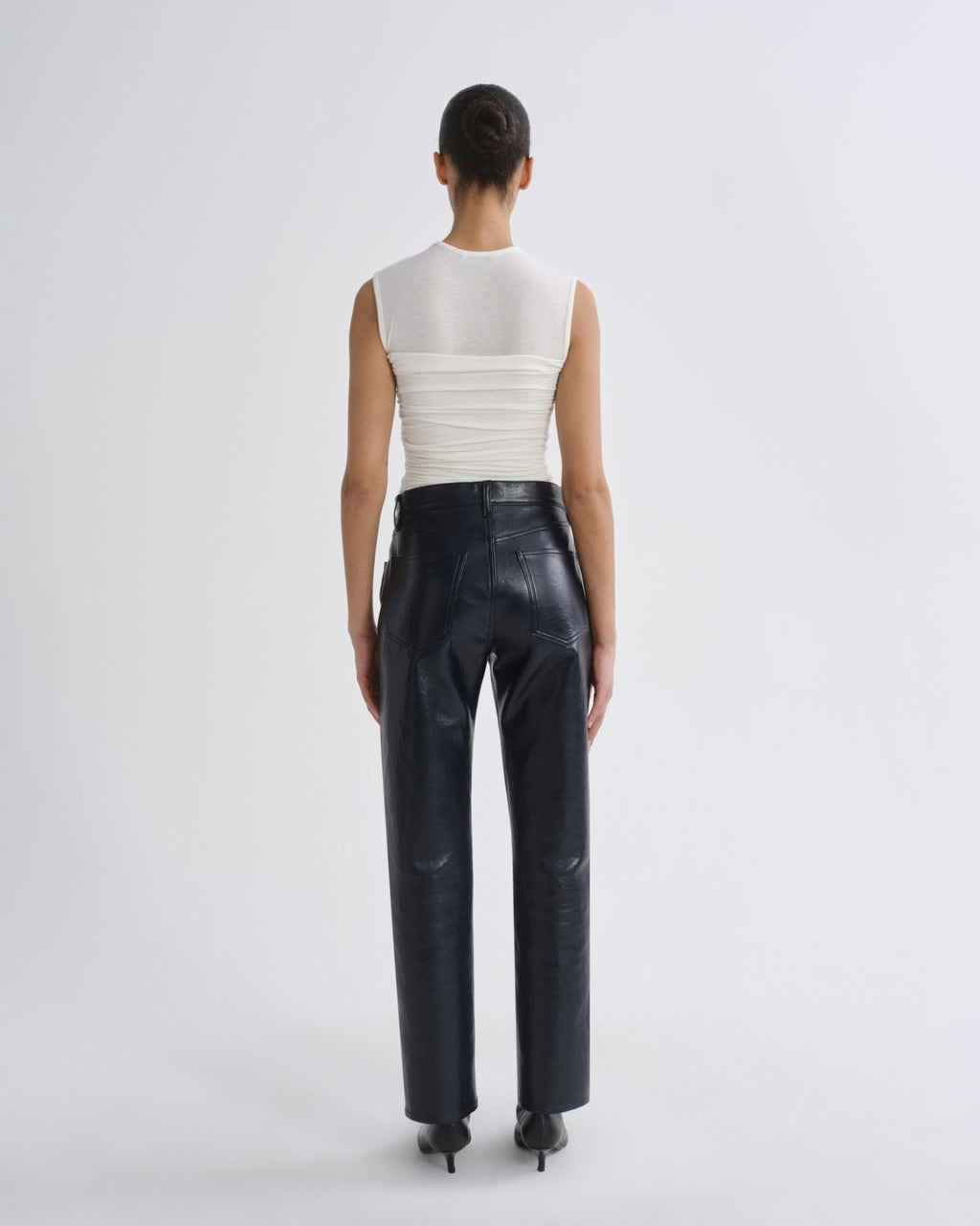 A model shown from the back wearing a white sleeveless ribbed top with a sheer upper back panel and dark navy wide-leg leather pants, set against a plain light gray studio background.