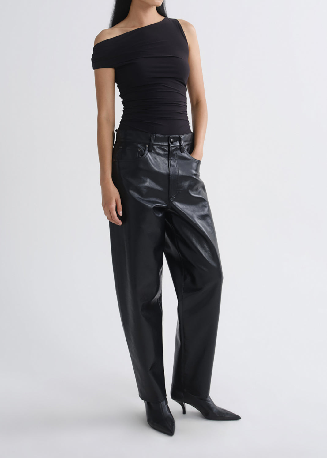 A model wearing a black off-the-shoulder ribbed top and shiny black wide-leg faux-leather pants with black pointed-toe heels, photographed against a grey background.