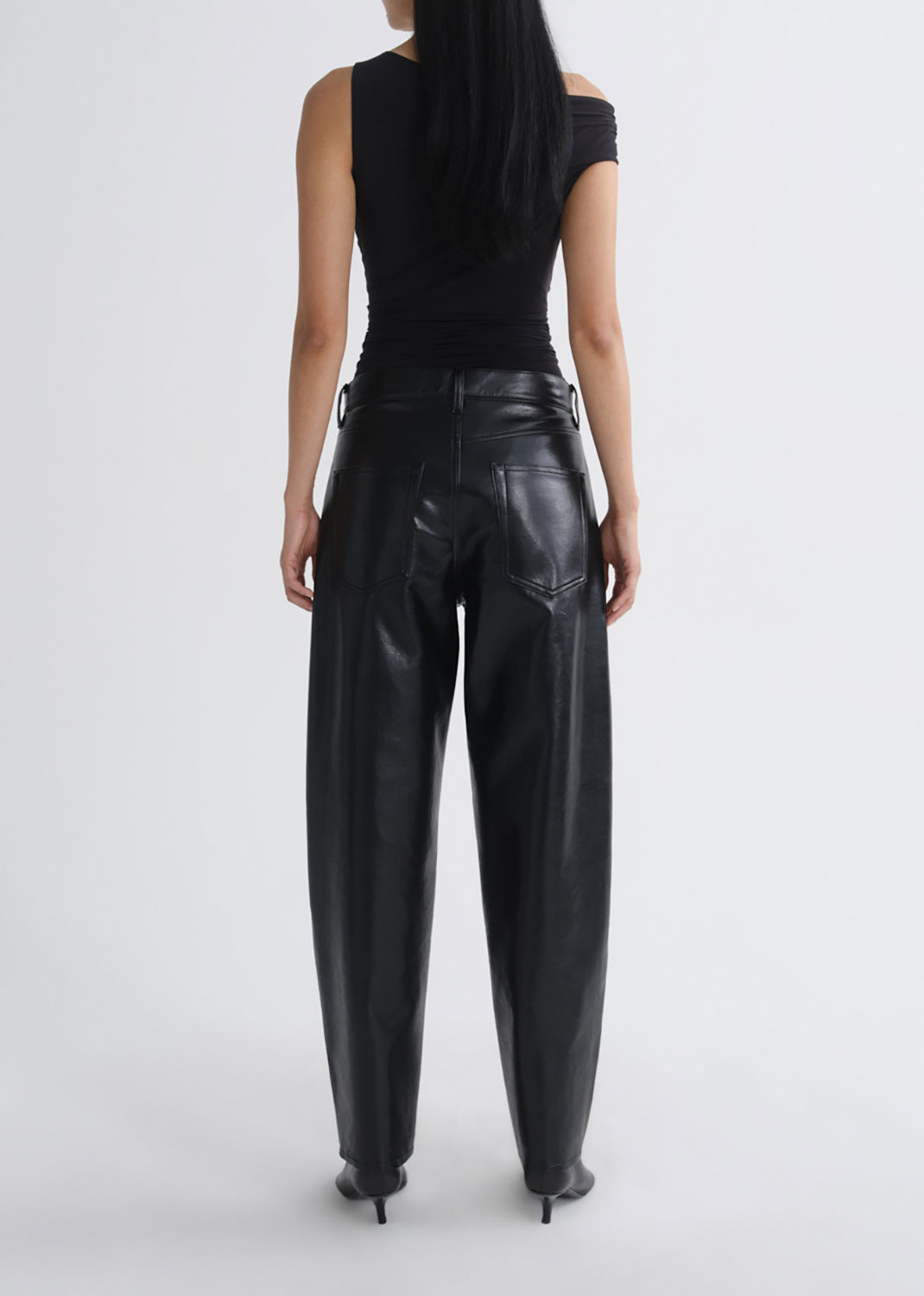 A model shown from the back wearing black wide-leg faux leather pants, a black off-the-shoulder top, and black pointed-toe heels against a grey background.