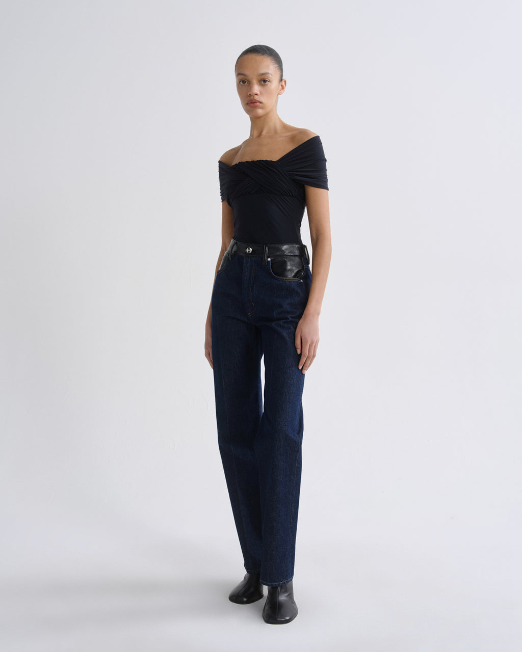 Woman wearing dark indigo straight leg jeans with a black off-the-shoulder top on a grey background.