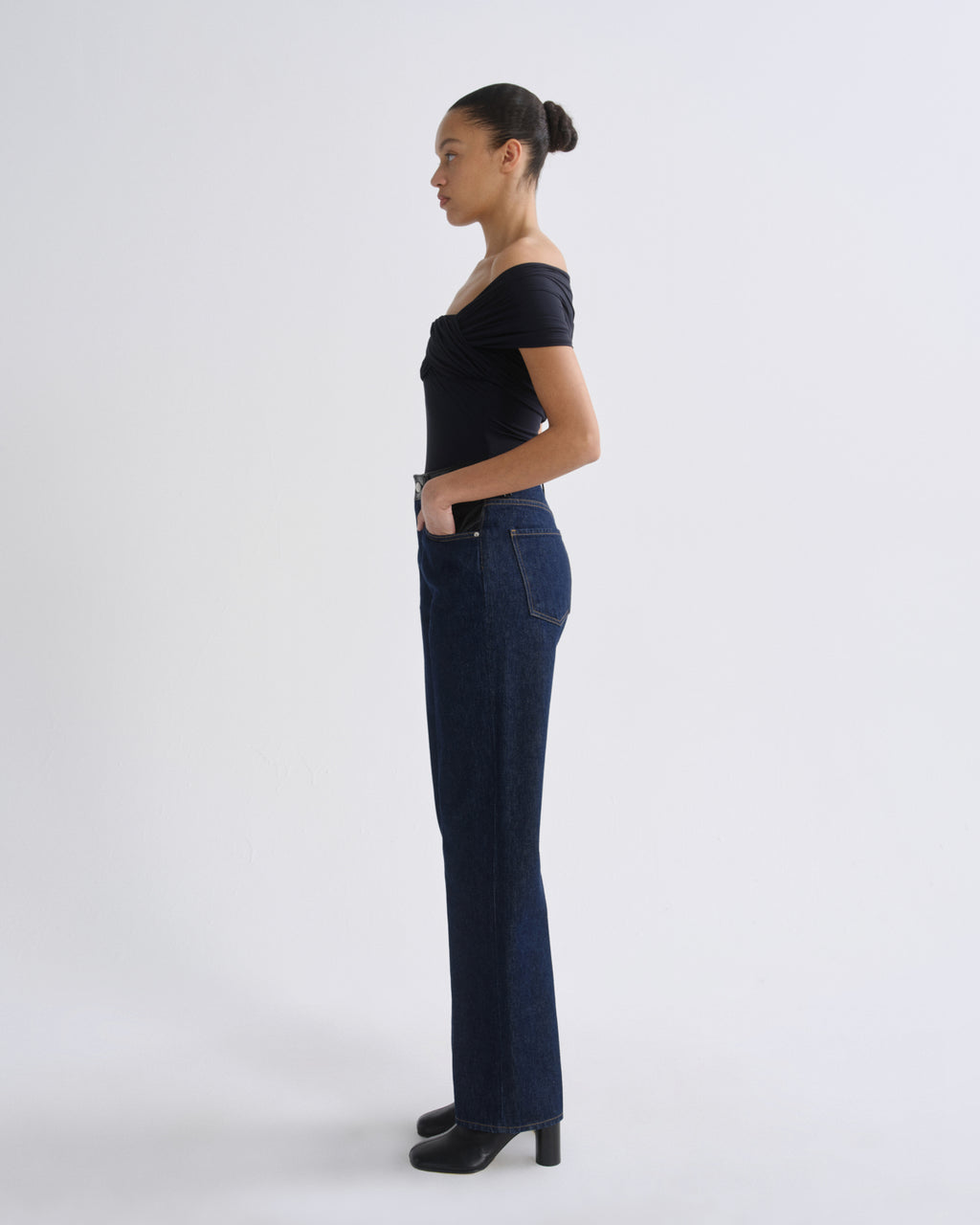 Woman wearing dark indigo straight leg jeans with a black off-the-shoulder top viewed from the side on a grey background.