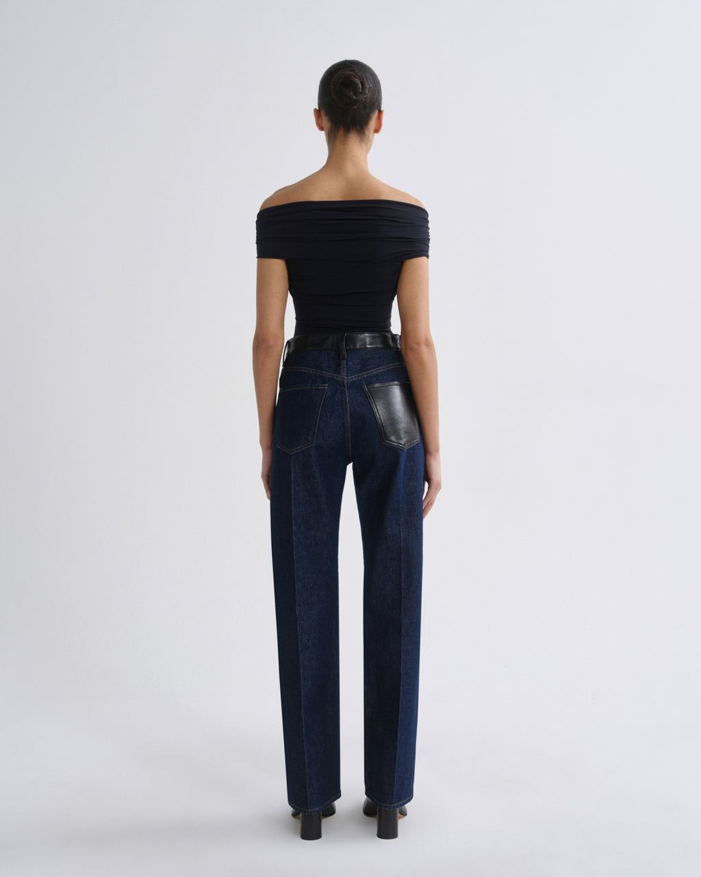 A model viewed from the back wearing dark blue high-waisted straight-leg jeans with black leather back pocket accents, paired with a black off-the-shoulder top and black heels, against a light gray studio background.
