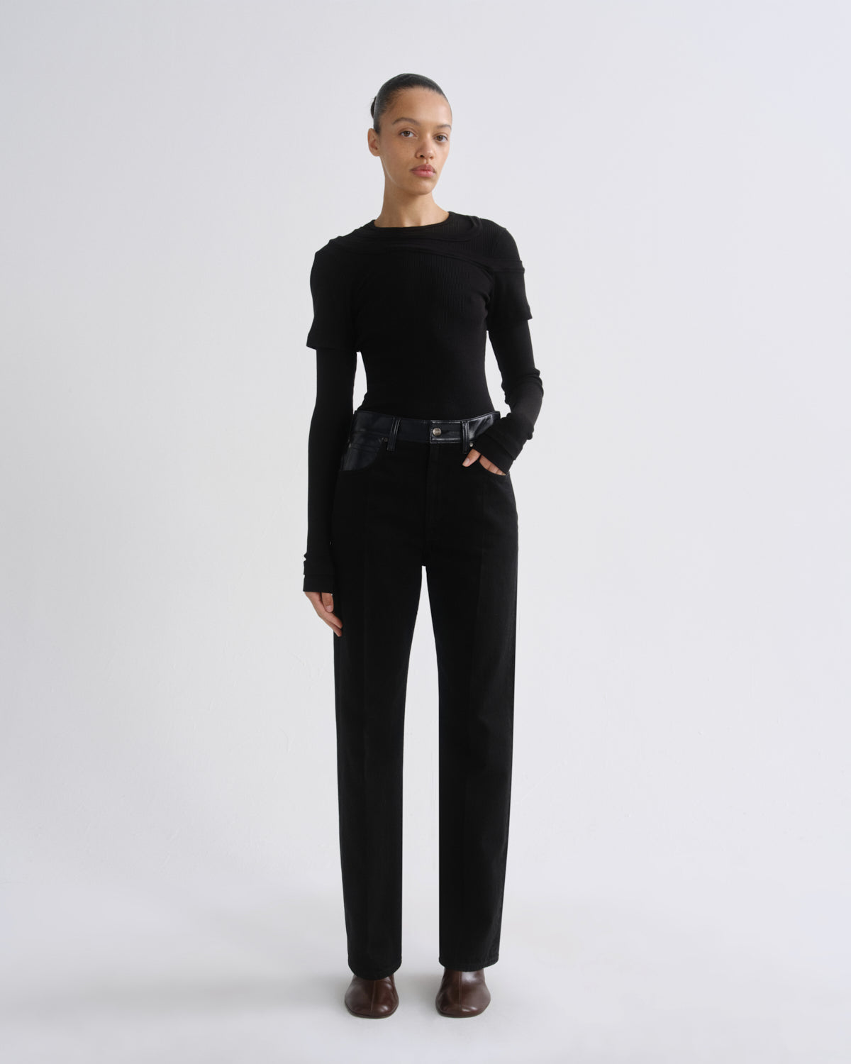 A model stands against a light gray studio background wearing a black long-sleeve top with textured shoulders and black wide-leg trousers, styled with a dark belt and brown shoes.